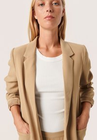 Béžový blazer s dobře padnoucím střihem, vyrobený z hladké tkaniny, s klopovým límcem, kapsami a ohrnutými rukávy přes bílý strukturovaný top.