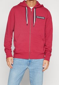 Haut zippé rouge en tissu doux, avec une poche avant, des cordons blancs et un badge logo sur la poitrine gauche.