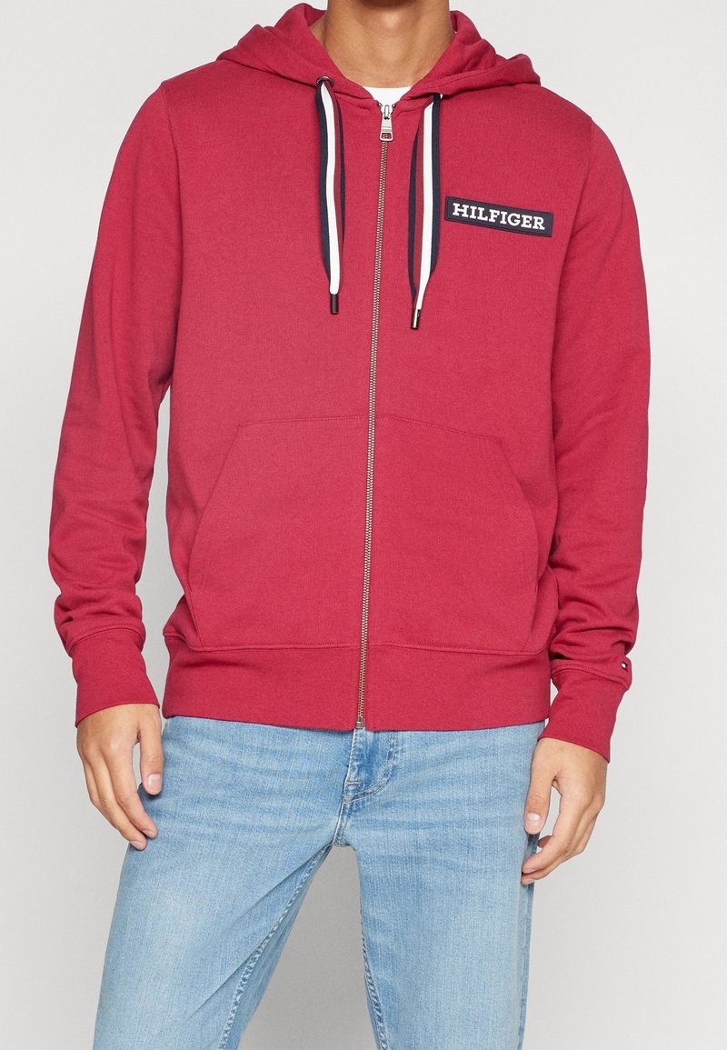 Haut zippé rouge en tissu doux, avec une poche avant, des cordons blancs et un badge logo sur la poitrine gauche.