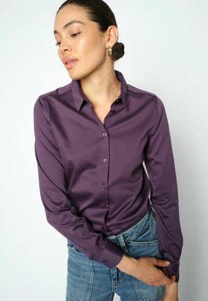 Woman with dark hair in a bun wearing gold hoop earrings, a purple button-up shirt, and blue jeans adjusting her sleeve against a white background.
