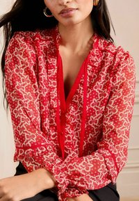 Blusa rossa a fantasia con dettagli arricciati, con scollatura a V profonda, chiusura a bottone e maniche lunghe con polsini arricciati. Tessuto semitrasparente.