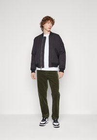 Giubbotto bomber nero con polsini a costine, indossato sopra una t-shirt bianca, abbinato a pantaloni in velluto a coste verde oliva e sneakers bianco e nero.