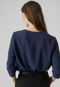 Navyblauwe langemouwen top met een gestructureerde afwerking, een ontspannen pasvorm, ronde halslijn en omgeslagen mouwen, gecombineerd met zwarte high-waisted broek.