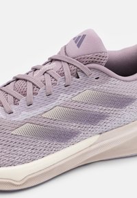 Zapatilla deportiva de color púrpura claro con parte superior de malla, superposiciones texturizadas y tres franjas en contraste. Suela curva suave con detalle de marca.