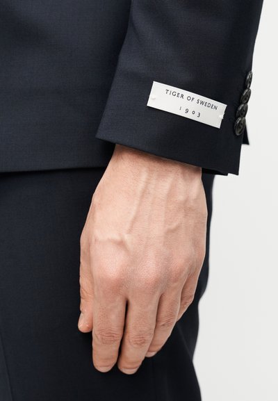 Μανίκι σακακιού ναυτικού μπλε με ετικέτα υφάσματος που αναγράφει "TIGER OF SWEDEN 1903" και τέσσερα μαύρα κουμπιά, που αναδεικνύουν την ομαλή υφή και την εφαρμογή ραφή.