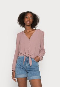 Blush long-sleeve blouse with a V-neck, button-down front, and tied hem, paired with rolled-up denim shorts. Soft texture and lightweight fabric.