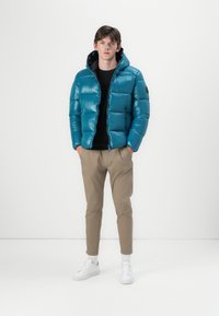 Teal puffer jacket with a hood and quilted design, worn over a black top. Paired with light brown pants and white sneakers.