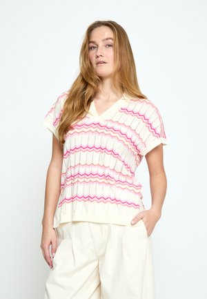 Jonge vrouw met lang blond haar, die een mouwloze trui met V-hals draagt met roze zigzagstrepen en crème wijde broek, staand met hand in de zak.