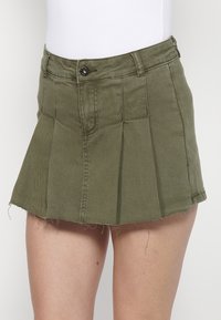 Mini-jupe en denim vert olive avec bouton devant, passants de ceinture, coutures et ourlet effiloché portée avec un haut blanc.
