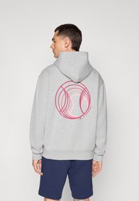 Grå hættetrøje med et stort pink cirkulært grafisk motiv på ryggen, der har koncentriske cirkler. Kængurulomme og justerbar hætte.