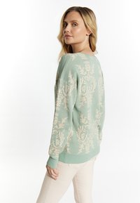 Hellgrüner Pullover mit cremefarbenem Blumenmuster, lockere Passform, weiter Ausschnitt und gerippte Bündchen. Kombiniert mit hellbeigen Hosen.