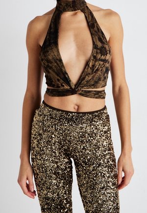 Mujer con un top halter marrón con estampado y escote en V pronunciado, y pantalones de lentejuelas doradas de talle alto, posando frente a un fondo liso.