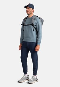 Mochila gris, sudadera azul claro, pantalones deportivos azul marino y zapatillas grises con detalles en negro. Atuendo deportivo casual, figura masculina de pie.