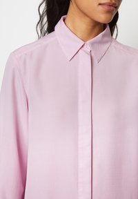 Camicetta rosa chiaro a maniche lunghe con colletto a punta, chiusura frontale con bottoni, texture liscia e dettagli di cucitura minimal.