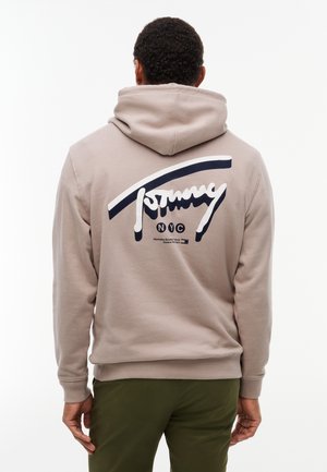 Mężczyzna w beżowej bluzie z kapturem z dużym granatowo-białym logo "Tommy NYC" na plecach, w połączeniu z oliwkowozielonymi spodniami, stoi tyłem.