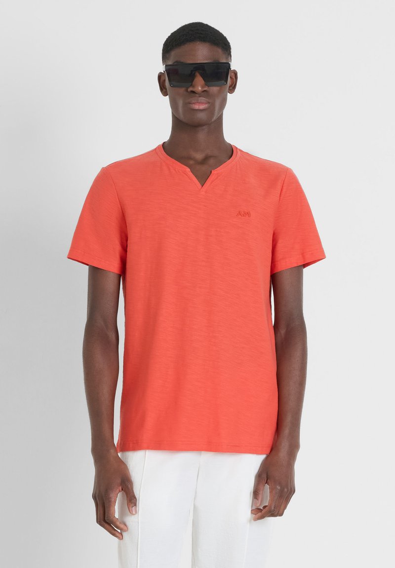 T-shirt a maniche corte, con scollo a V, di un vivace corallo. Realizzato in tessuto leggero con una leggera trama, presenta un piccolo logo in rilievo sul petto.