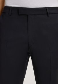 Schwarze maßgeschneiderte Hosen aus einem glatten Stoff, mit flachem Vorderteil, Gürtelschlaufen und einer subtilen Textur. Keine sichtbaren Taschen.