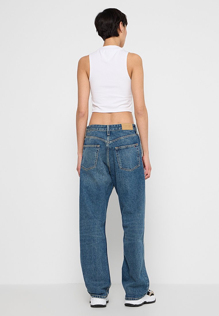 Osoba ubrana w białą bezrękawnikową bluzkę cropped, luźne niebieskie dżinsy i białe trampki, stoi przodem do gładkiej, jasnoszarej ściany.