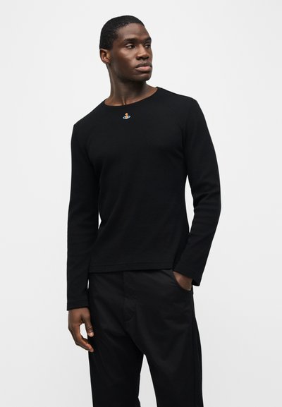 Άνδρας που φοράει μαύρο πουκάμισο με μακριά μανίκια και μικρό πολύχρωμο κεντητό λογότυπο στο στήθος, και μαύρο παντελόνι, στέκεται με το ένα χέρι στην τσέπη.