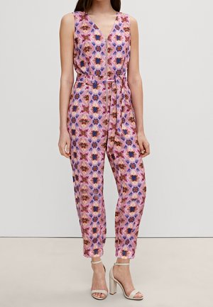 Combinaison rose sans manches avec motif floral bleu et marron, fermeture éclair à l'avant, ceinture nouée à la taille, portée avec des sandales à talons hauts beiges.