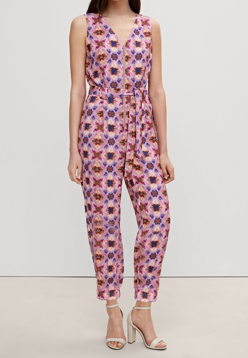 Combinaison rose sans manches avec motif floral bleu et marron, fermeture éclair à l'avant, ceinture nouée à la taille, portée avec des sandales à talons hauts beiges.
