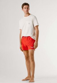 Jonge man die blootsvoets staat, gekleed in een wit T-shirt en rode zwemshorts met het logo "Pepe Jeans" op het linkerbeen.