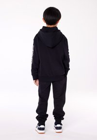 Felpa nera con cappuccio e pantaloni coordinati, con polsi a coste. Sneakers con accenti bianchi e blu navy completano l'outfit.