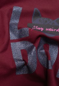 Maroen stof met grote, donkergrauwe glinsterende letters die "L O D" spellen en een silhouet van een kat. De tekst luidt "Blijf vreemd" in het roze. Zachte textuur.