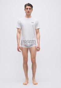 Νέος άνδρας στέκεται ξυπόλητος φορώντας ένα λευκό T-shirt Moschino με λογότυπο στο τελείωμα και εφαρμοστά λευκά εσώρουχα boxer.