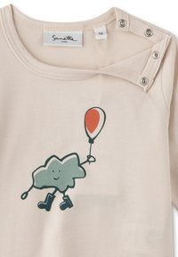 Cremefarbenshirt mit rundem Ausschnitt, das einen grünen Wolkencharakter zeigt, der einen rot-weißen Ballon hält, und schwarzen Stiefeln. Druckknopfdetail an der Schulter.
