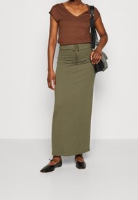 Camiseta marrón de cuello en V combinada con una falda verde oliva de longitud a la rodilla con cintura ajustable, acentuada por unos zapatos planos negros y un bolso.