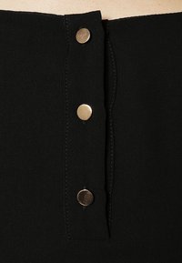 Svart tyg med en vertikal rad av fem cirkulära knappar i guldton, med sydda sömmar och en slät yta.