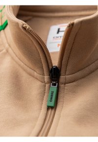 Giacca beige con zip e colletto alto; texture morbida; presenta una zip nera con un tirante verde etichettato "OMBRE."