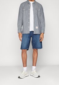 Hombre con zapatillas blancas, calcetines blancos, pantalones cortos de mezclilla azules, camiseta blanca y una camisa de rayas grises abotonada, de pie contra un fondo blanco.