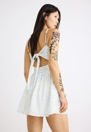 Pitkätukkainen nainen, jolla on valkoinen kuvioitu mekko, jossa on avoin selkä ja jonka takana on rusetti, näyttää kukkataideteoksia käsivarressaan.