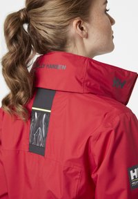 Rode waterdichte jas met een hoge kraag, contrasterend zwart paneel met een plastic zak en reflecterende accenten. Logo op de kraag.