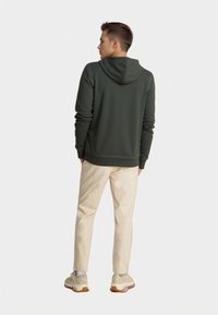 Felpa con cappuccio verde scuro, dal tessuto liscio, con polsini e orlo elasticizzati, abbinata a pantaloni beige chiaro e sneakers dai toni neutri.
