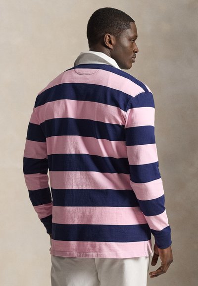 Mann trägt ein Rugbyshirt mit langen Ärmeln mit breiten horizontalen dunkelblauen und rosa Streifen, weißem Kragen und weißen Hosen, seitlich.