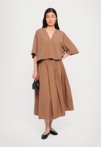 Brun cropped topp med V-hals, kombinert med et langt, fullt brunt skjørt. Svart håndveske og flate sko fullfører antrekket. Glatt stofftekstur.