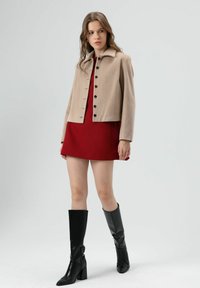 Beige cropped jack met een kraag, zwarte knopen, gecombineerd met een rode jurk en zwarte kniehoge laarzen met hak, staand tegen een neutrale achtergrond.