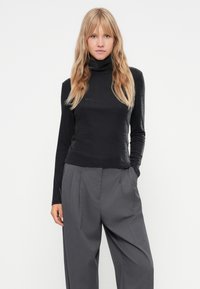 Μαύρο, εφαρμοστό πουλόβερ με ψηλό γιακά και μακριά μανίκια, κατασκευασμένο από γυαλιστερό υλικό. Συνδυάζεται με γκρι παντελόνια ψηλής μέσης που έχουν χαλαρή εφαρμογή.