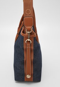 Denim axelväska med bruna läderdetaljer, guldhårdvara och ett strukturerat band. Har en framträdande logotyp och kontrasterande sömmar.