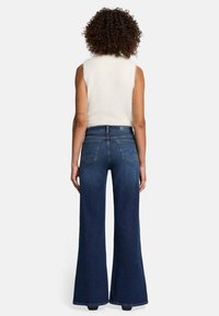 Jeans in denim blu scuro a zampa d'elefante con tasche posteriori, indossati con un top bianco senza maniche. Presentano una vita alta e una finitura testurizzata.