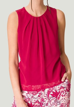 Ärmellose, leuchtend pinke Bluse mit Falten am Ausschnitt, kombiniert mit weißen und pinken Hosen mit Blumenmuster.