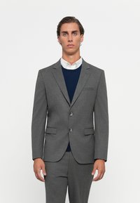 Grau Anzugjacke mit zwei Knöpfen, Reverskragen und seitlichen Taschen, getragen über einem marineblauen Pullover. Glatter Stoff mit taillierter Passform.