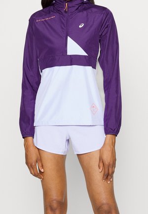 Femme portant une veste de sport à manches longues violette et lavande claire avec un short assorti sur un fond uni.