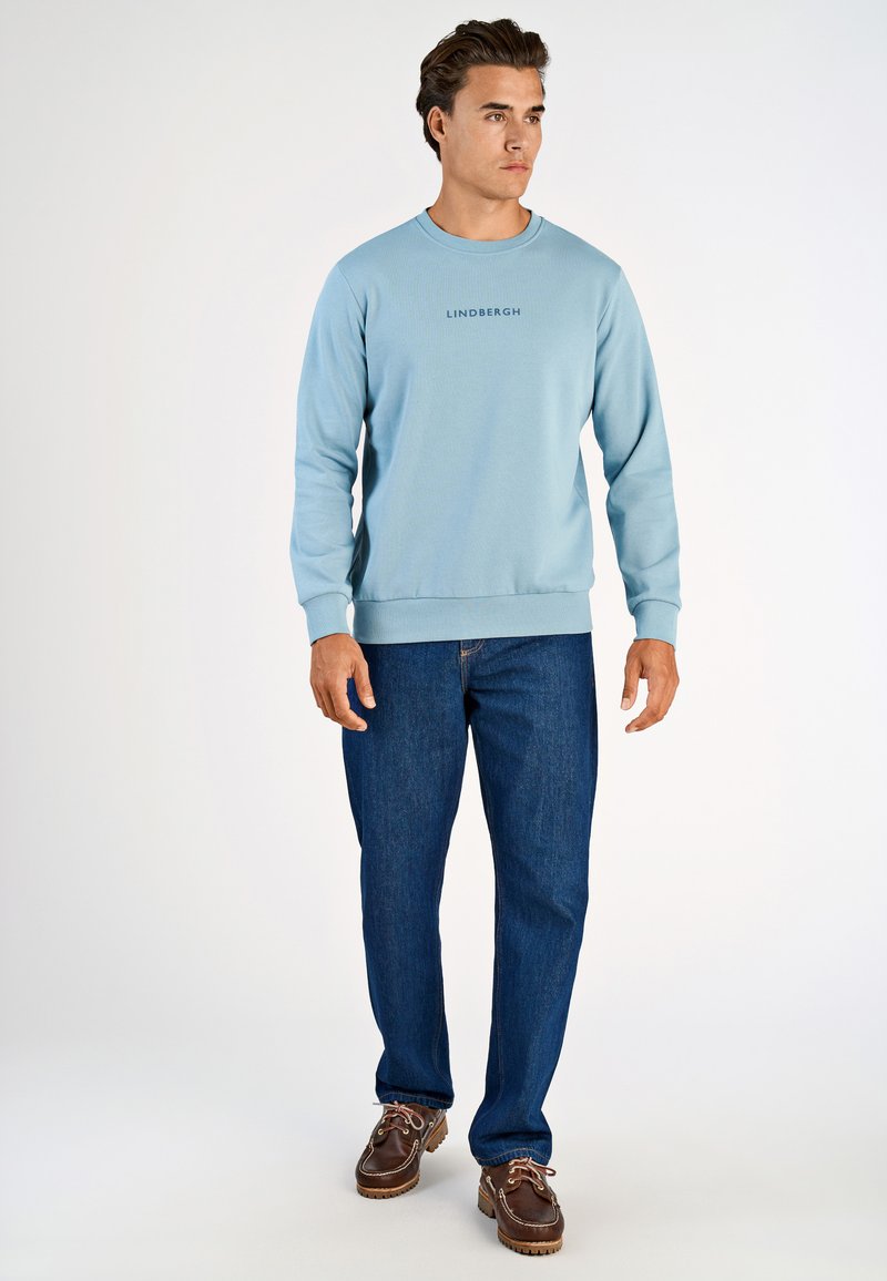 Ljusblå sweatshirt med rundhals och ribbade muddar, med texten "LINDENBERGH". Kombinerad med blå jeans och bruna skor.