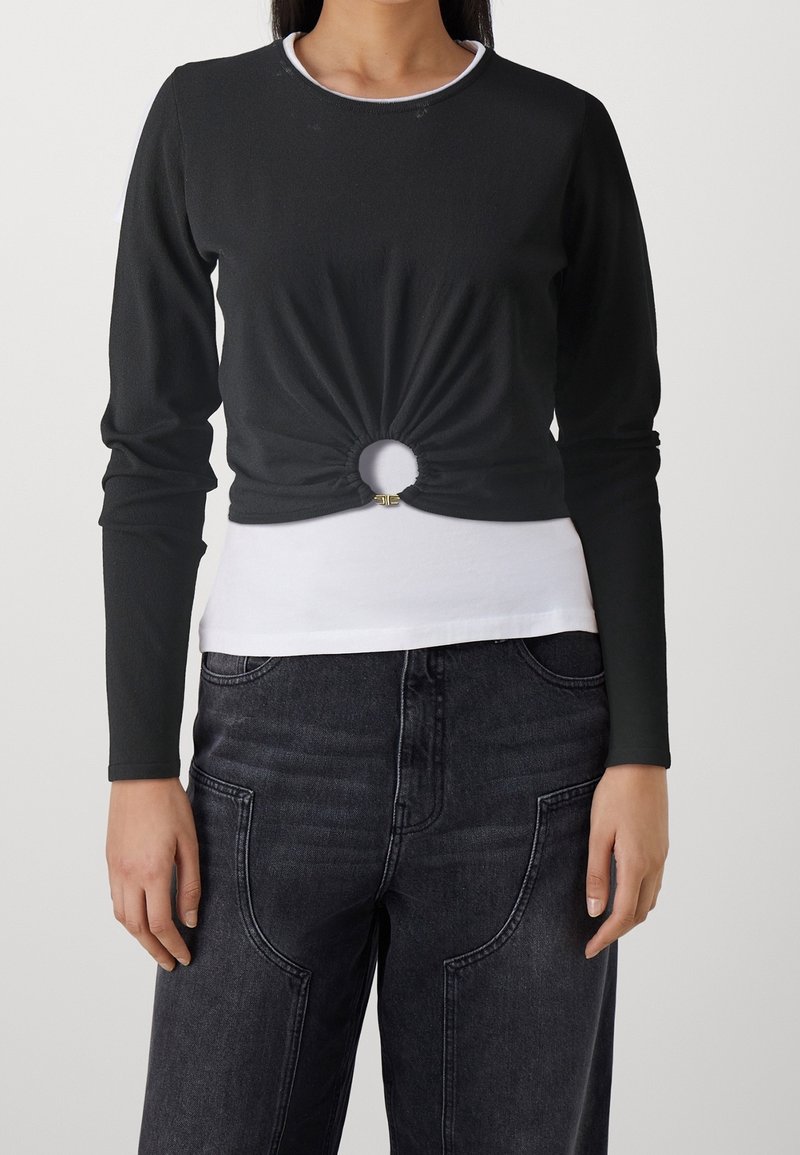 Femme portant un crop top noir à manches longues avec une découpe circulaire et une petite boucle au-dessus d'un sous-pull blanc et un jean en denim foncé.