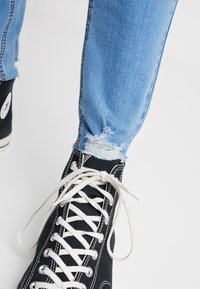 Zapatillas altas negras con cordones y ojales blancos, combinadas con unos vaqueros desgastados de color azul claro con dobladillos deshilachados.