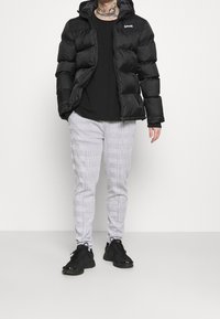 Schwarze Pufferjacke mit Kapuze, Reißverschluss und Logo-Patch; kombiniert mit grauen karierten Hosen und schwarzen Sneakers.